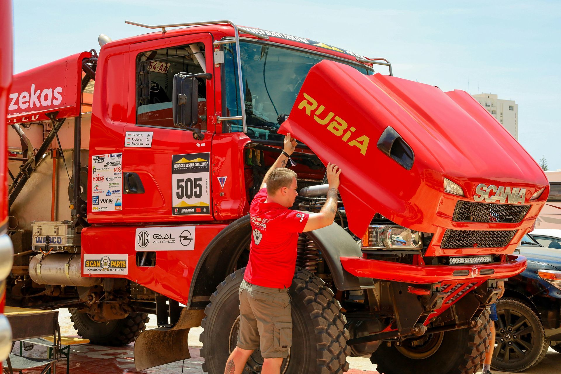 Man in red shirt opens hood of red Scania truck, labeled