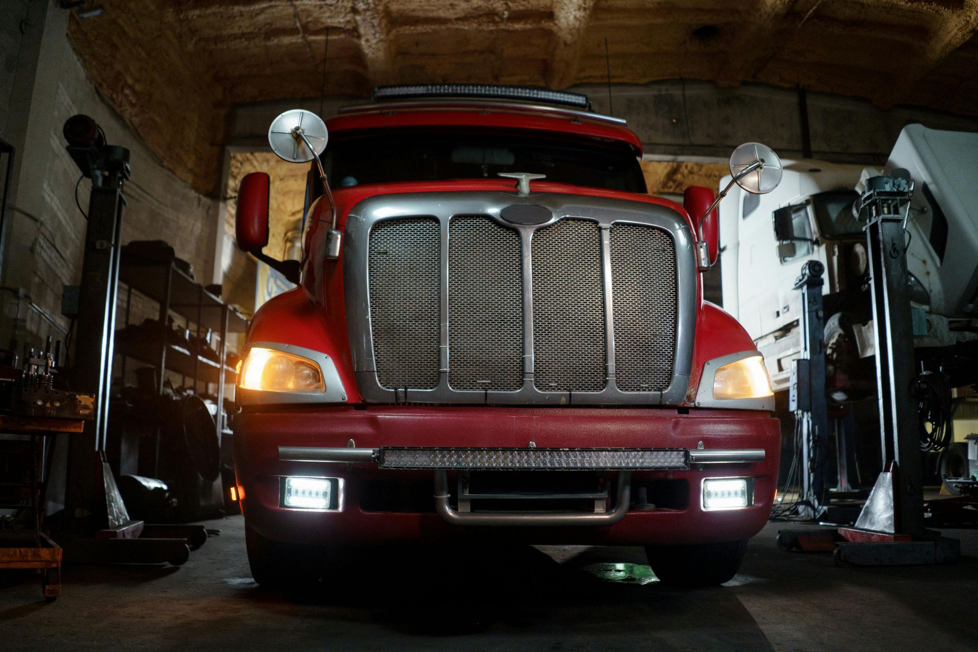 Red semi-truck parked inside a dimly lit garage. Headlights on, surrounded by tools and equipment.