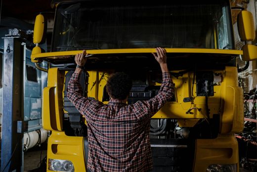 Mechanic in plaid shirt working on the front of a yellow truck in a garage.