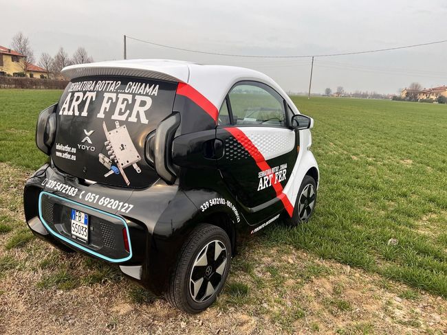 Una piccola auto bianca e nera con una grafica vistosa parcheggiata su un prato sotto un cielo nuvoloso.