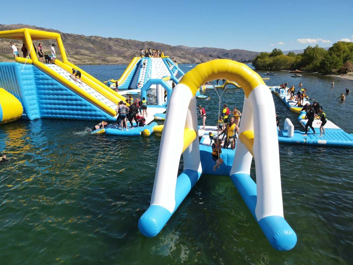 Children and adults having fun on colorful inflatable slides and obstacles at a lake waterpark on a sunny day.