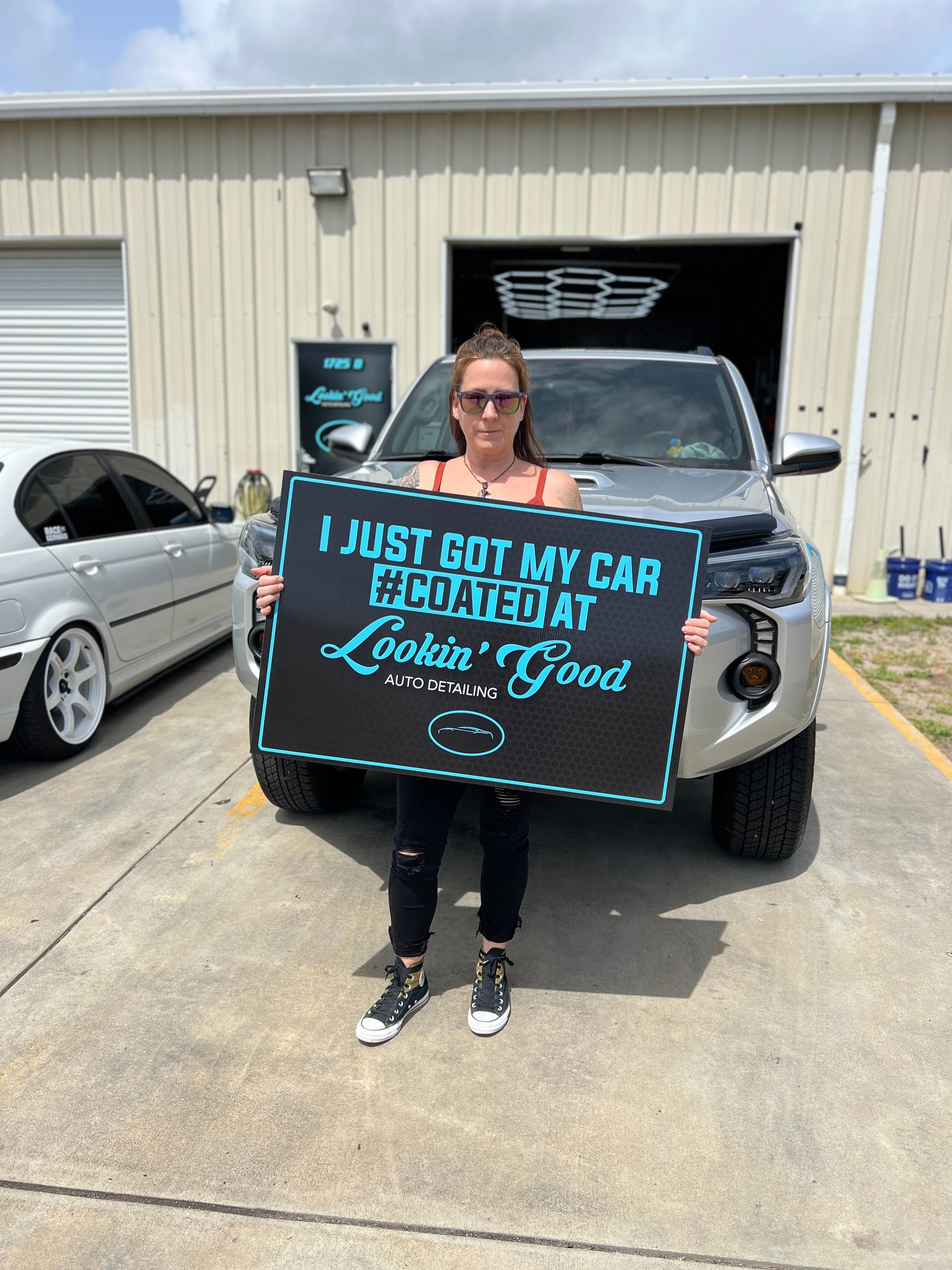 Woman with sign in front of a truck outside auto shop. The sign says 