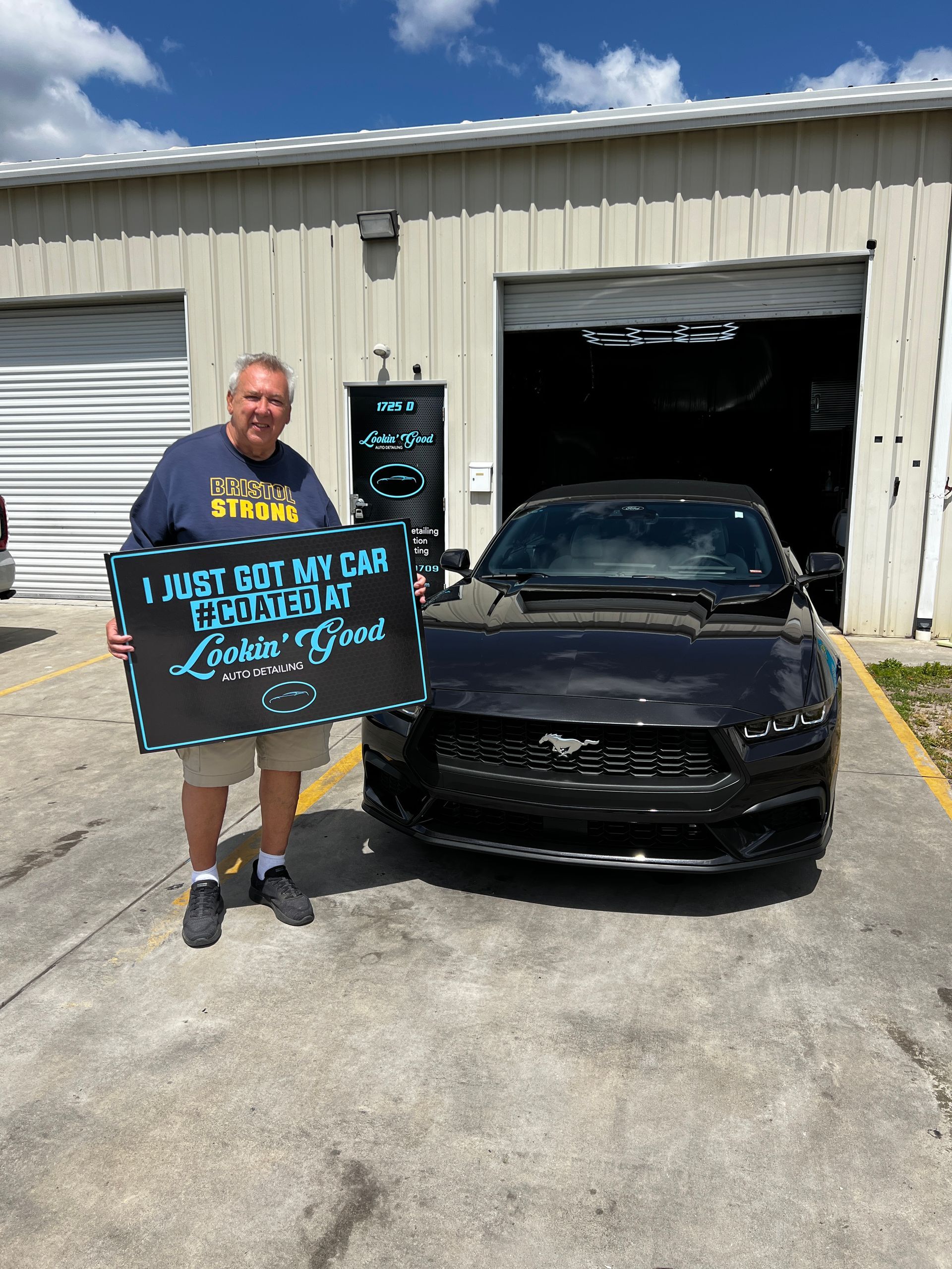 Man holding sign next to black Mustang in front of a garage. The sign reads 