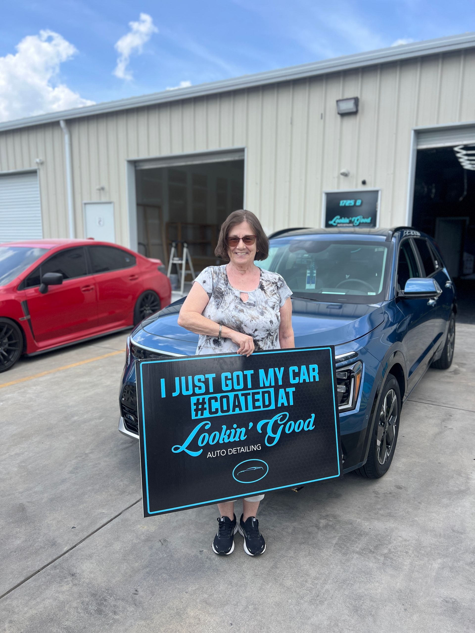 Woman smiling, standing in front of a blue SUV, holding a sign: 