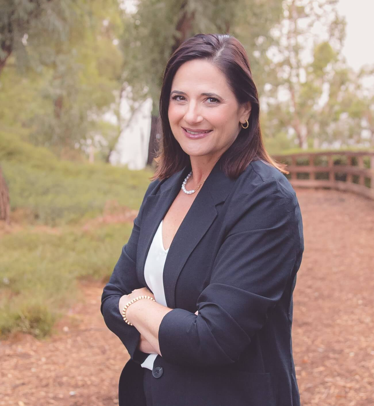 A woman in a black jacket is standing in a park with her arms crossed.