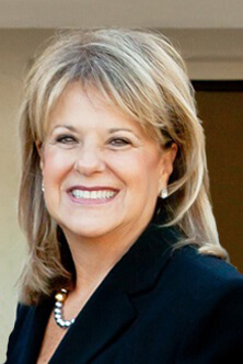 A woman in a black jacket and pearl necklace is smiling.