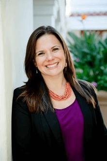 A woman in a black jacket and purple shirt is smiling while leaning against a wall.