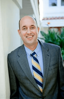 A man in a suit and tie is leaning against a wall and smiling.