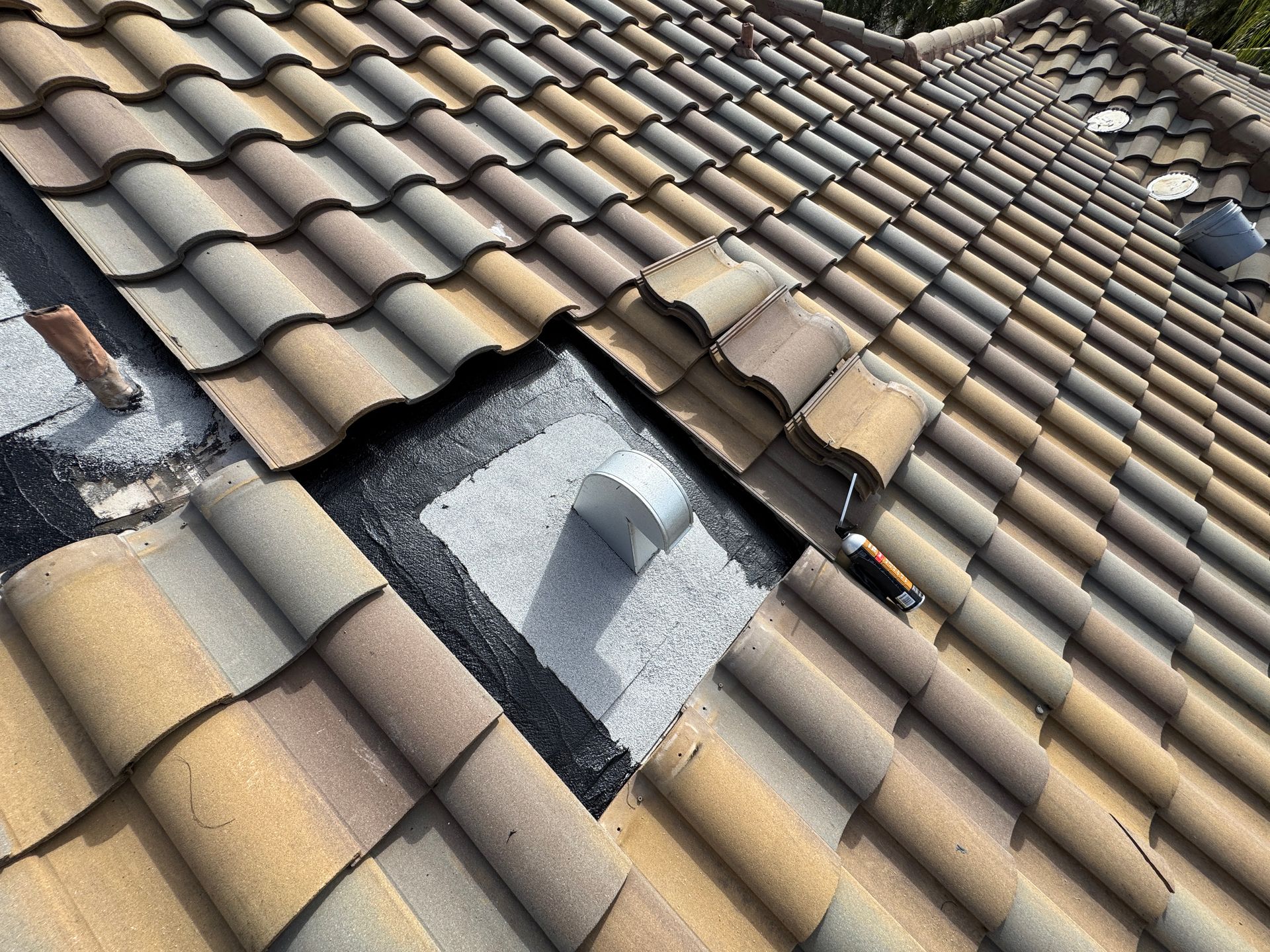 Overhead view of a clay tile roof with a square opening exposing the underlayment and a metal vent pipe.