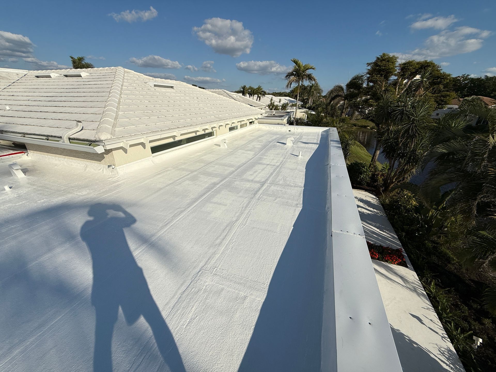 White roof with long shadows, visible person's shadow, blue sky with puffy clouds, trees.