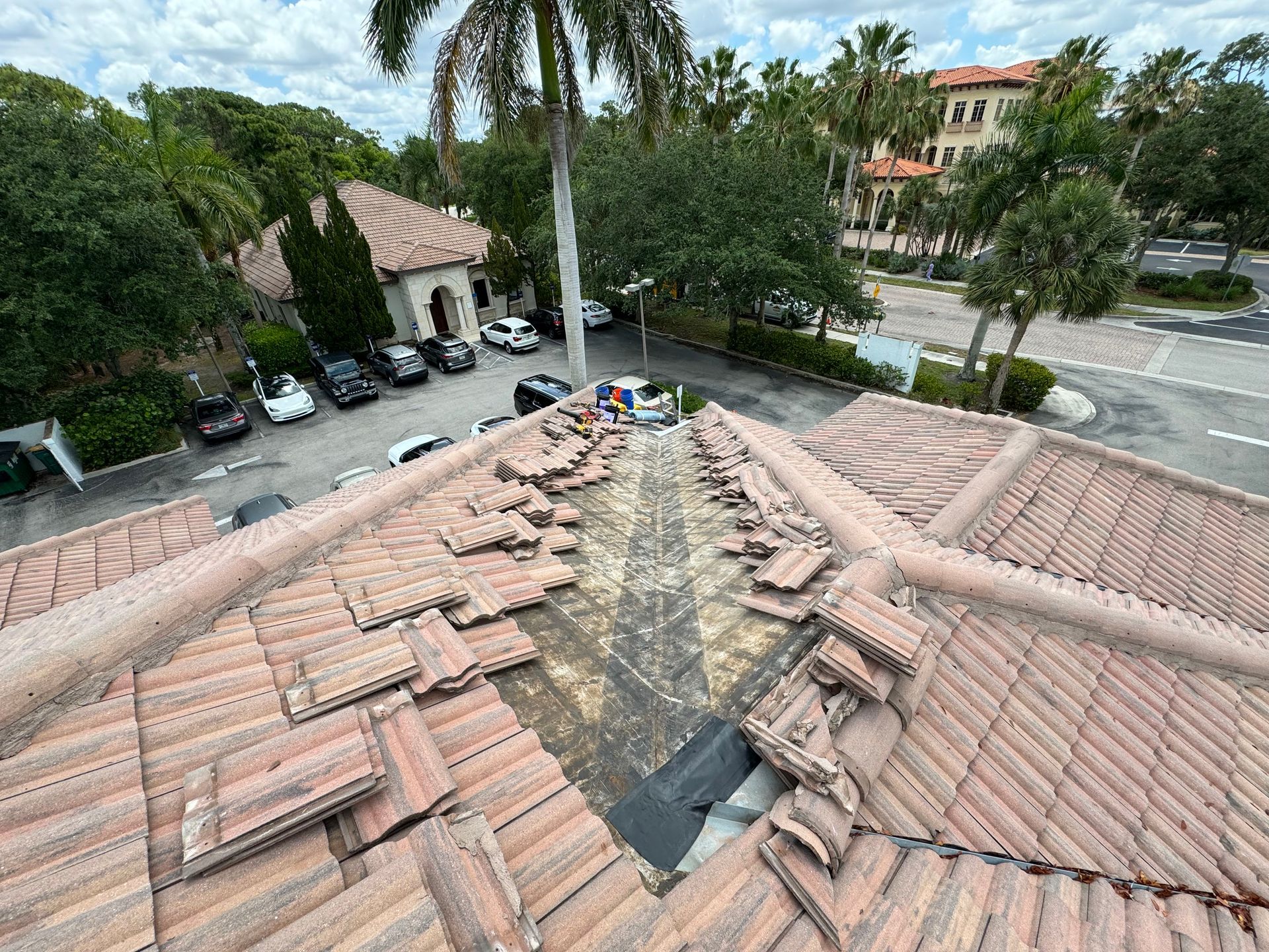 Roof tiles removed, exposing underlayment. Workers near a building with parked cars, palm trees, and blue sky.