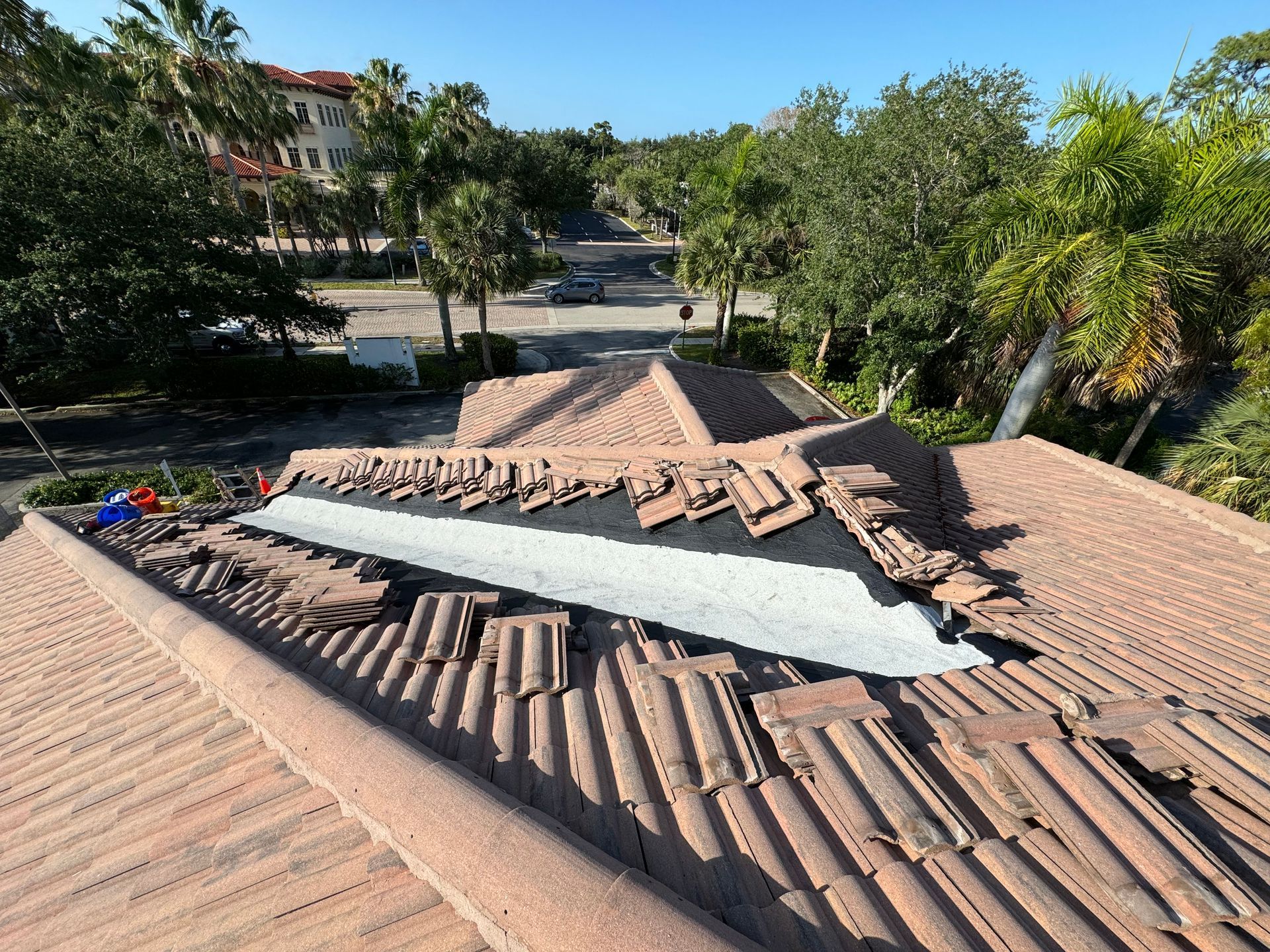 Rooftop with red tiles undergoing repair; silver flashing exposed, surrounded by broken tiles; palm trees in background.