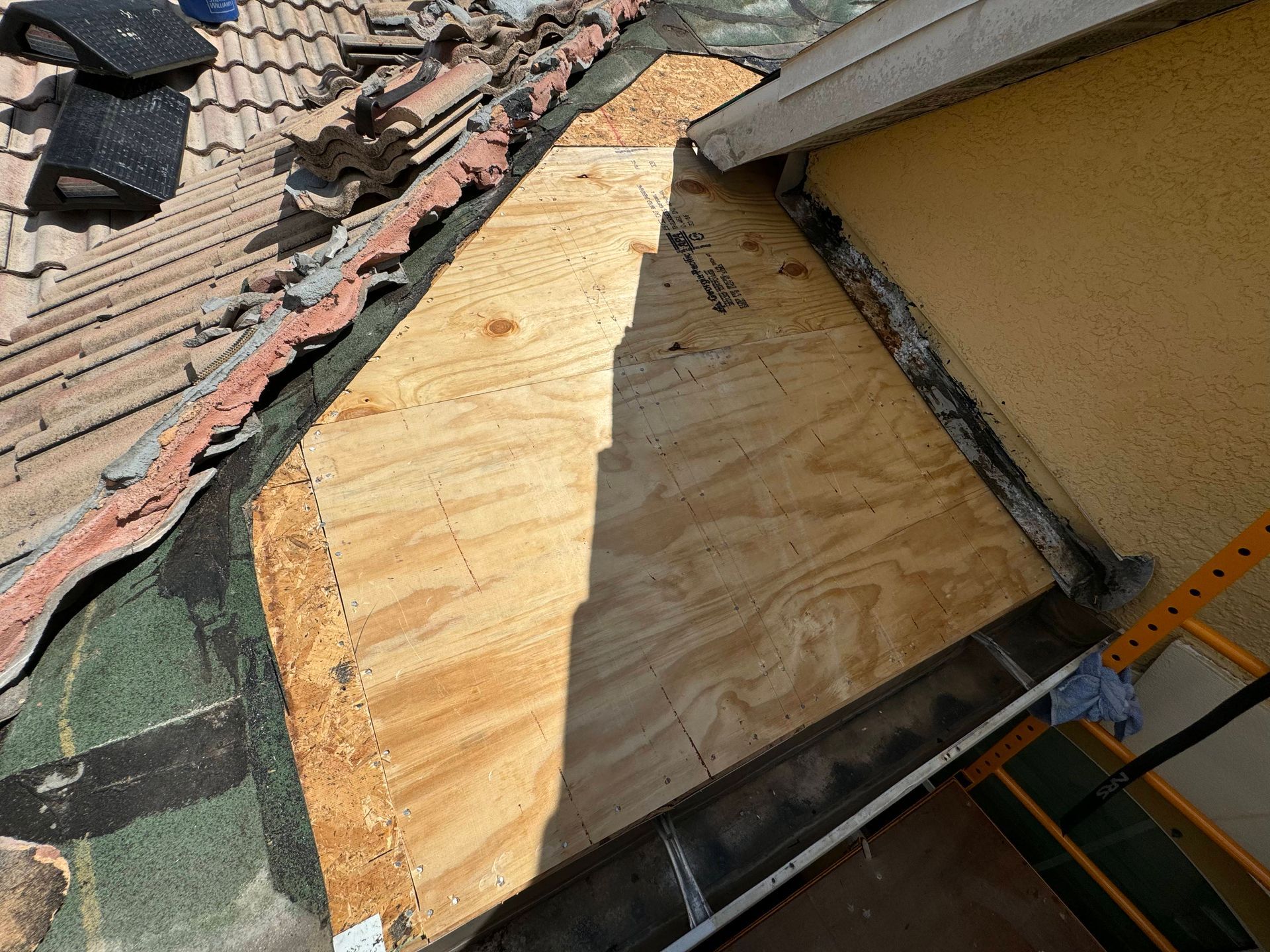 Roof section with plywood repair, partial terracotta tiles, black sealant, and stucco wall.