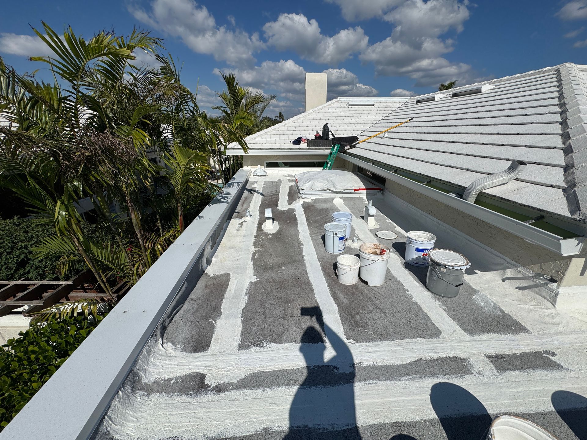 Flat roof being worked on, with white sealant applied in strips. Buckets and tools on the roof under a blue sky.