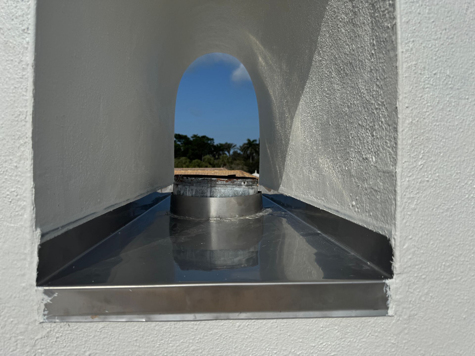 Looking through an arched window, a stainless steel drain is visible, with a view of trees and blue sky.