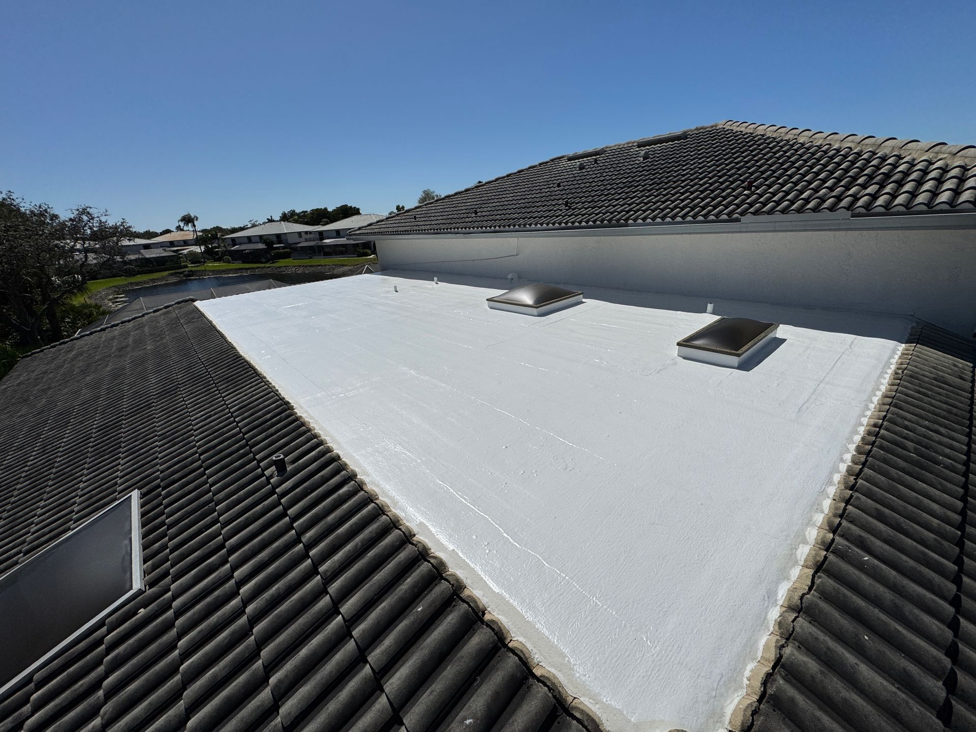 A roof with dark tiles bordering a white, flat section with two skylights, under a clear blue sky.