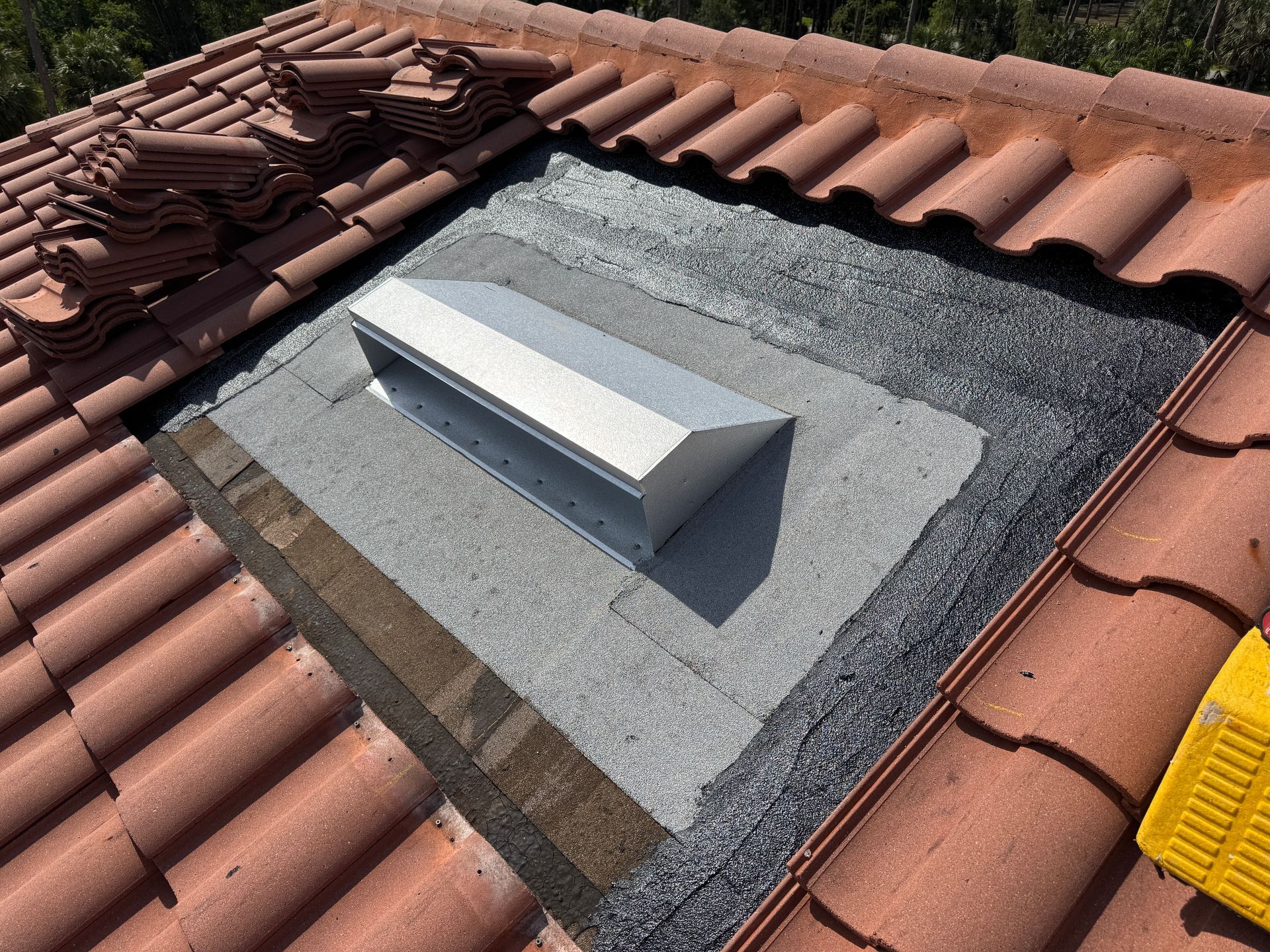 Roof vent surrounded by asphalt and clay tiles, with a stack of tiles in the background.