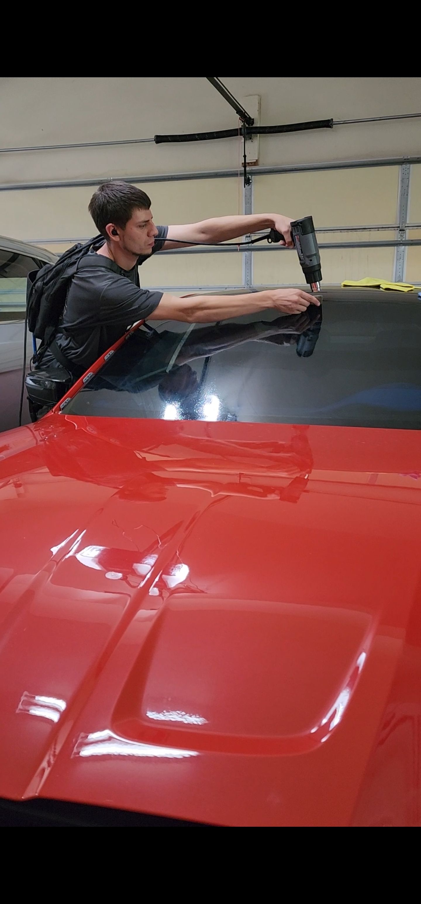 A man tinting a red car's window in a garage; he holds a tool as he works on the window.
