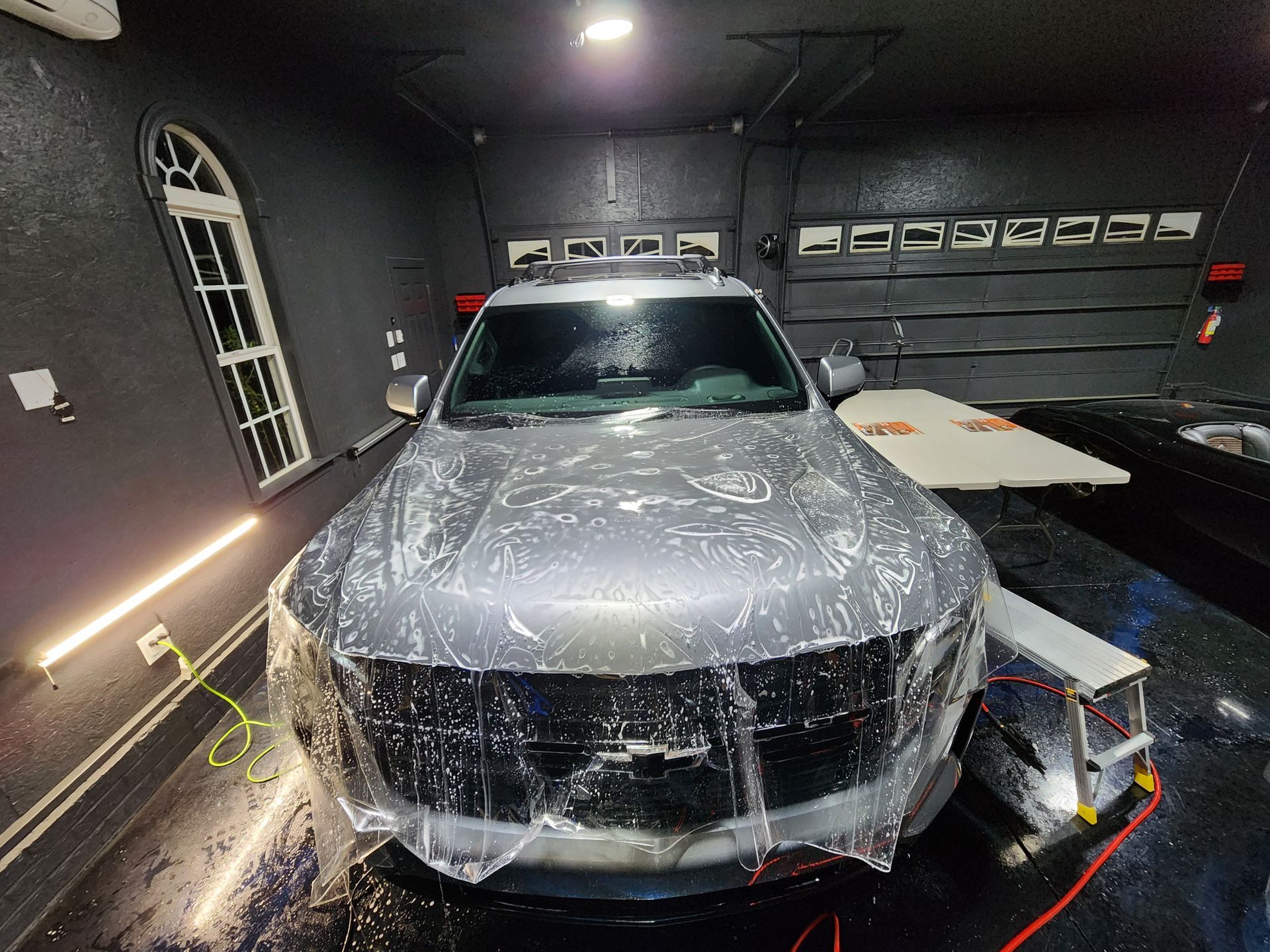 Silver car with plastic wrap on front in a car detailing bay.