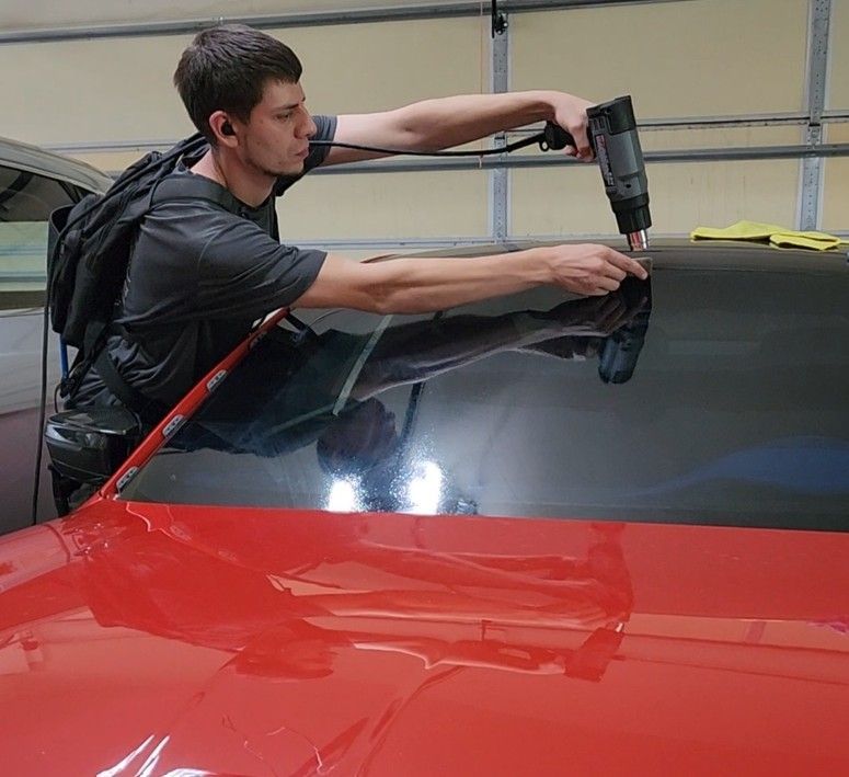Man applying window tint to a red car, using a heat gun. Indoors.