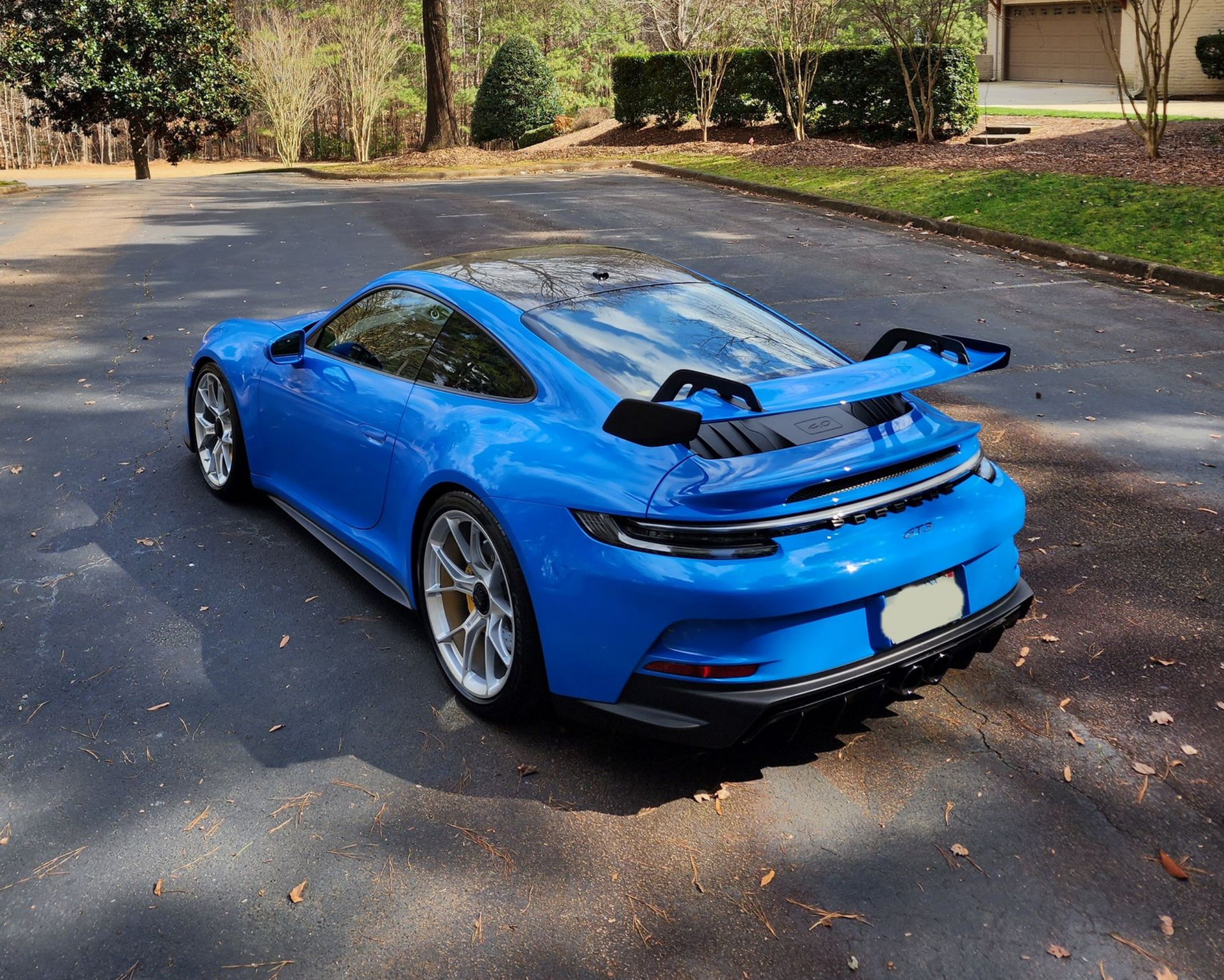 Blue Porsche sports car parked on a road with trees and a house in the background. Sunny day.