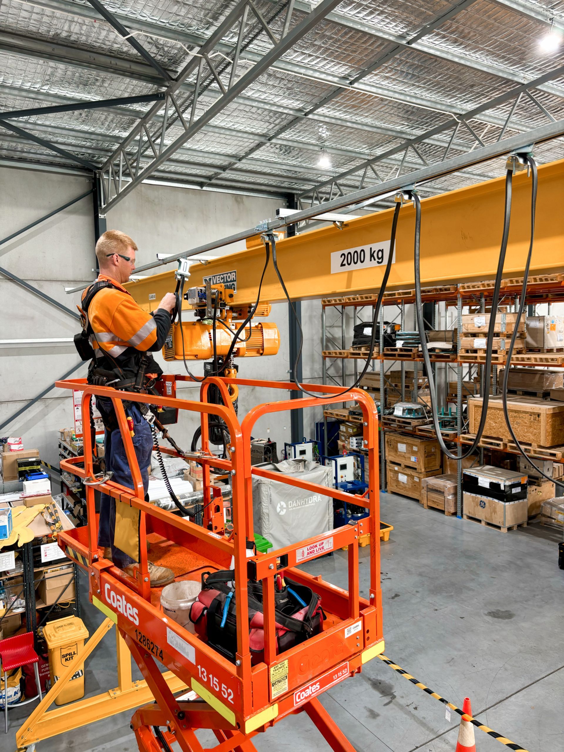 An Electrician is standing on a scissor lift working on a crane in a warehouse doing commercial electrical work.