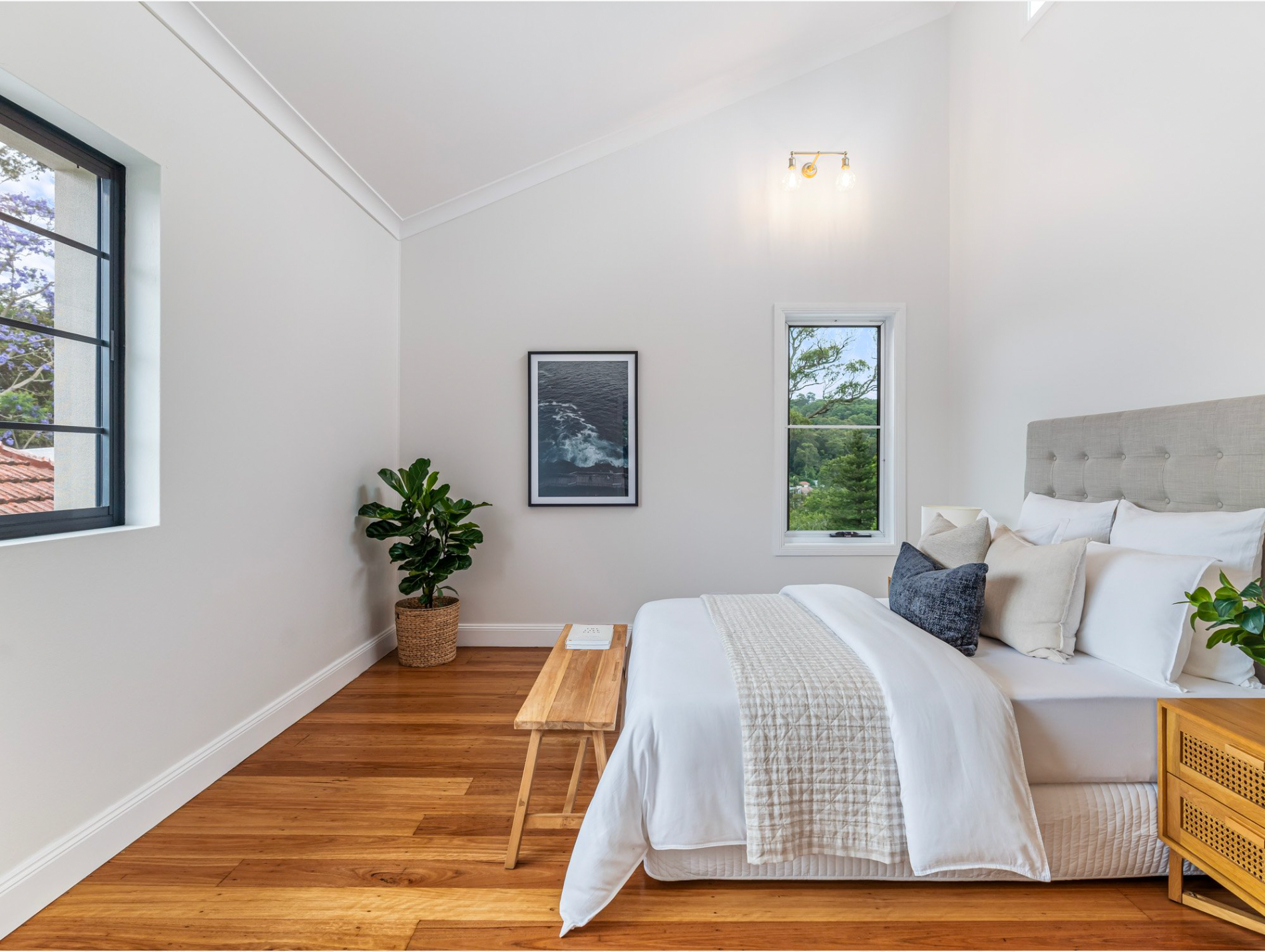 A bedroom with a bed , nightstand , bench, feature lighting and two windows.