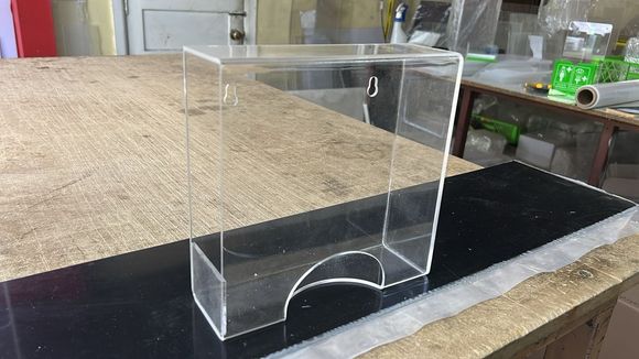 Un dispensador de cajas de acrílico transparente para montaje en pared, con un recorte curvo en la parte inferior, colocado sobre un banco de trabajo.