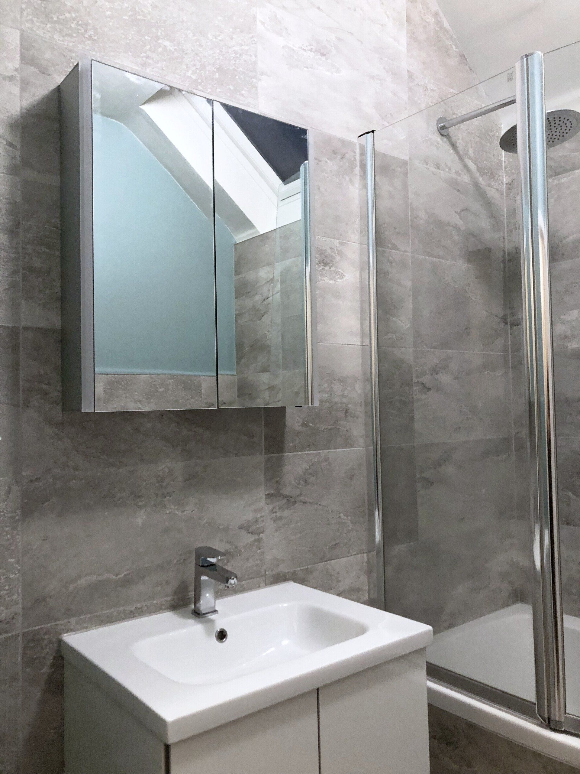A bathroom with a sink, mirror and shower and grey marble style tiles.