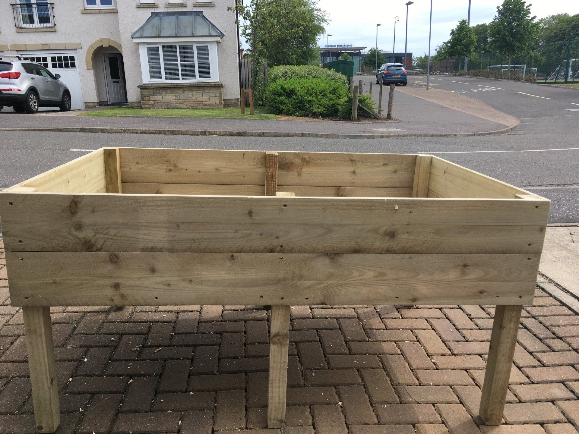 A wooden planter is sitting on a monoblock driveway in front of a house.