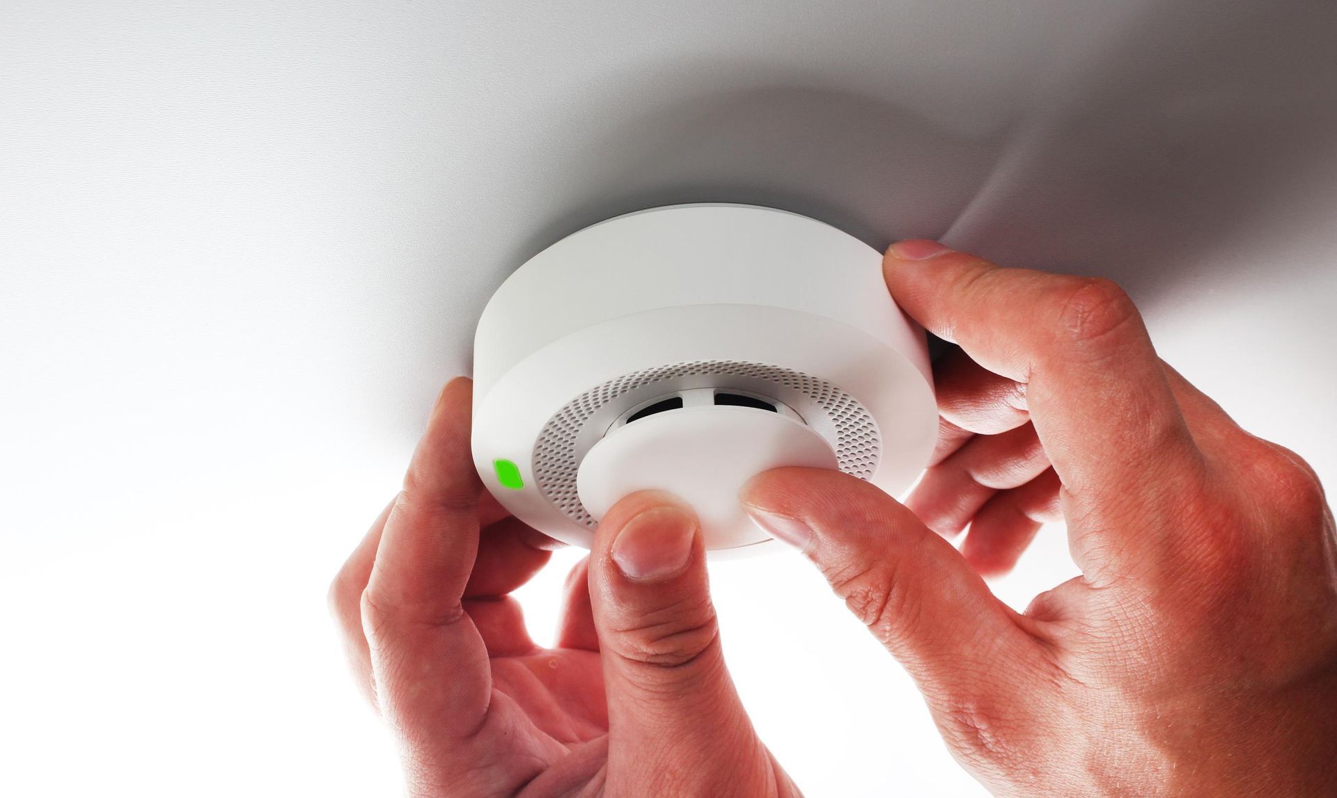 Hands installing a white smoke detector on a white ceiling.