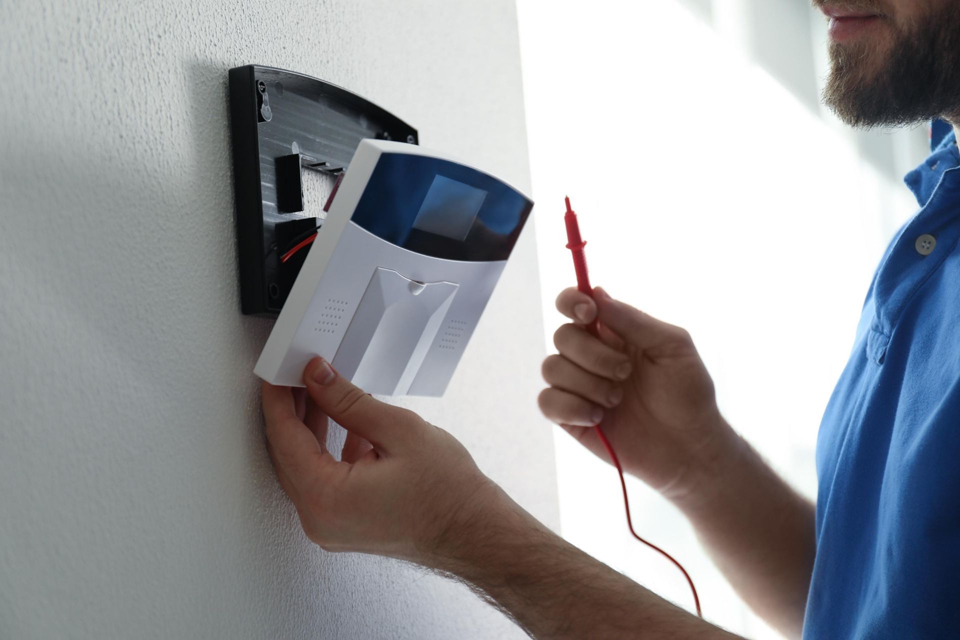 Person installing a security system control panel on a white wall, using a testing tool.