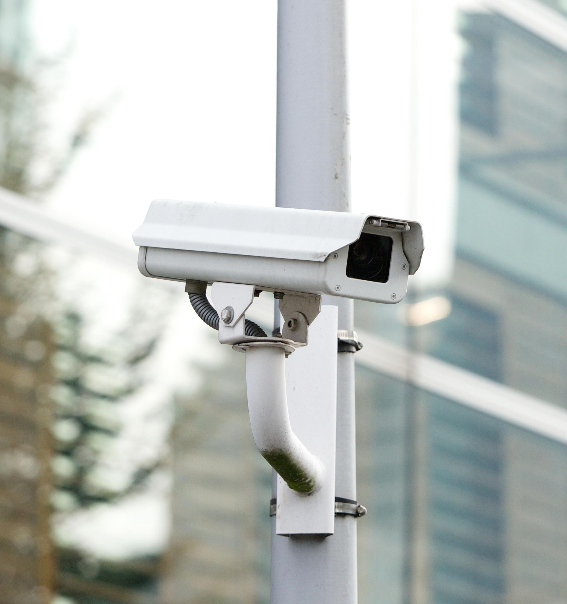 White security camera mounted on a metal pole, angled towards a glass building.