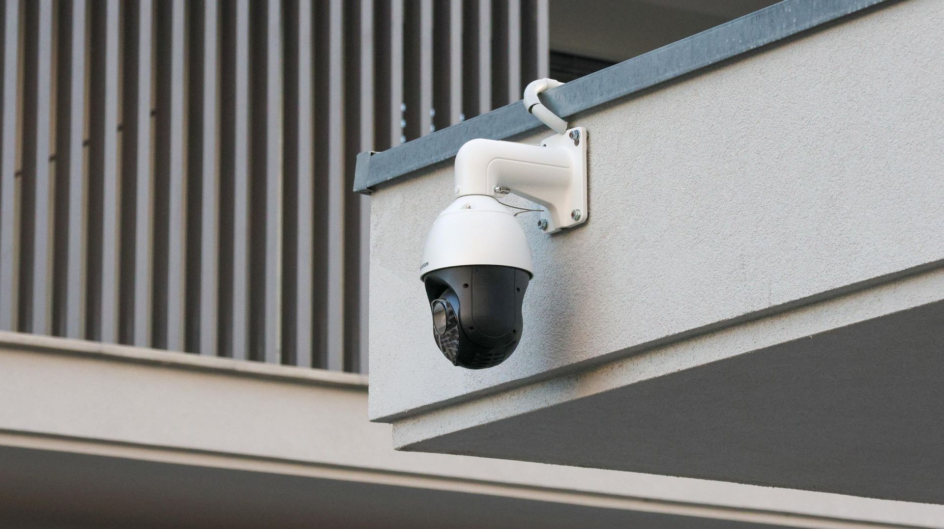 Security camera mounted on a building exterior, angled down. White camera, black dome, gray wall.