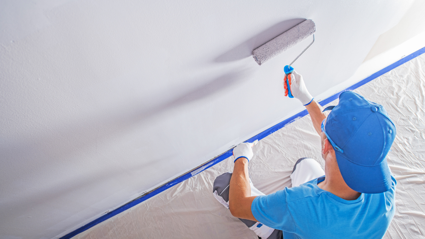 Person painting a white ceiling with a paint roller; blue painter's tape and clothing; soft lighting.