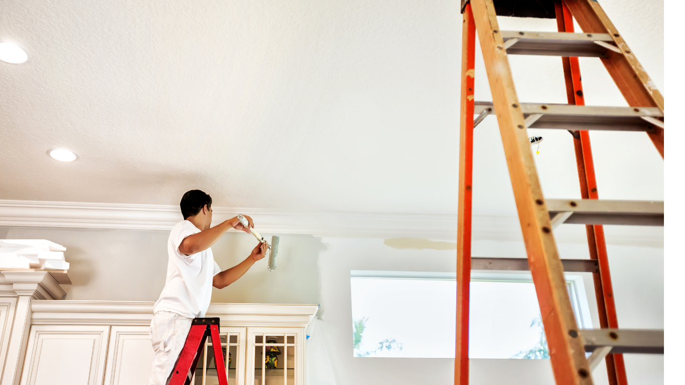 Person painting a ceiling standing on a red ladder in a room with white cabinets.