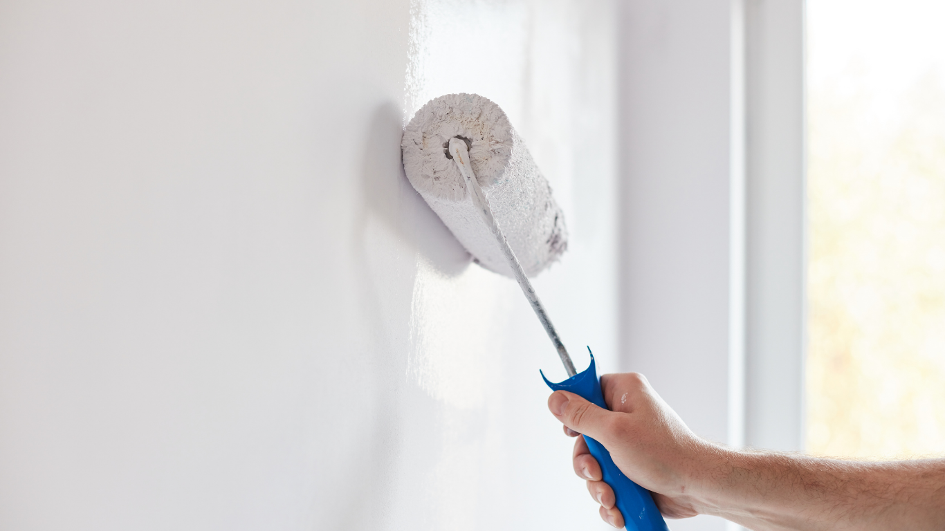 Hand using a paint roller on a white wall, near a window.