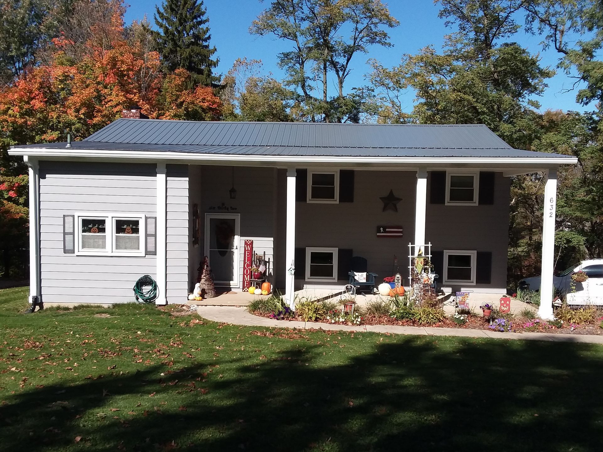 A single-story gray house with a metal roof and a front porch, surrounded by trees with autumn foliage.
