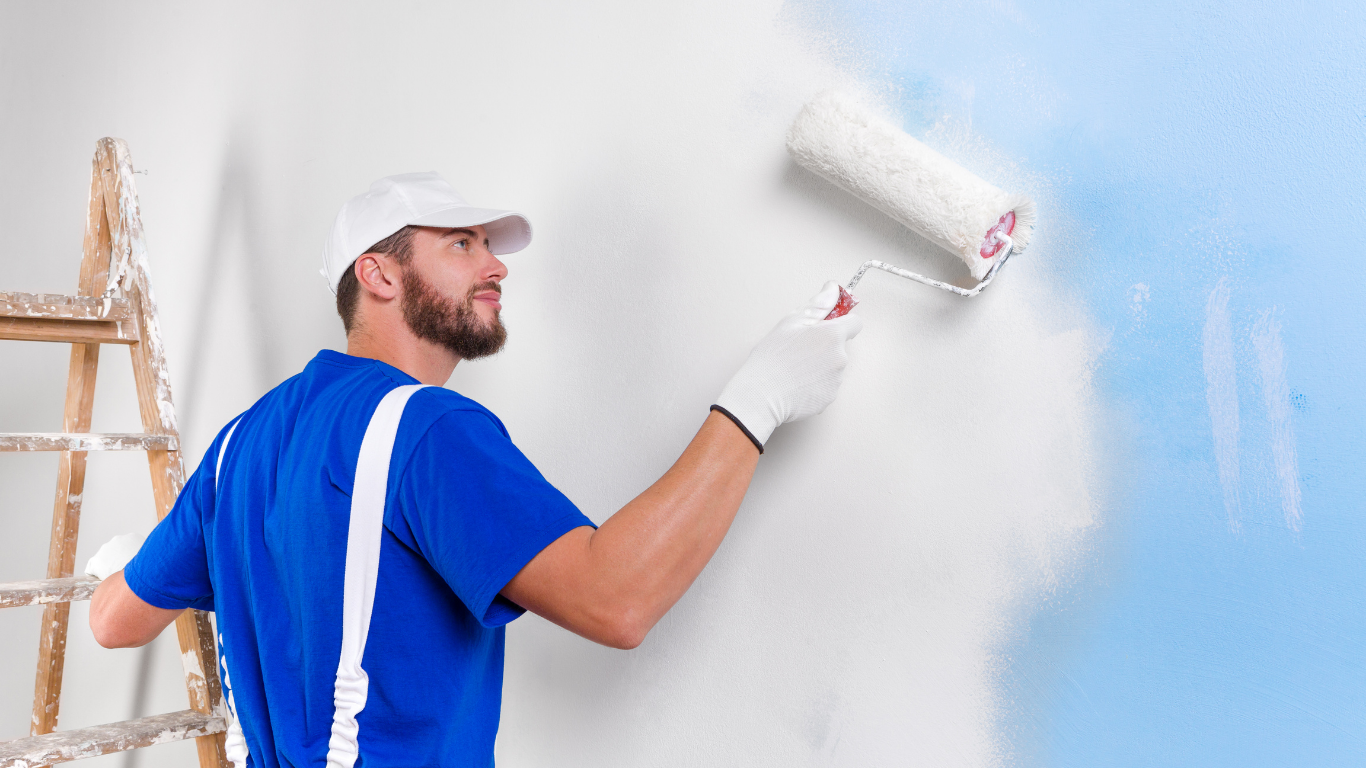 Person in blue overalls painting a wall white with a roller. Wooden ladder nearby.