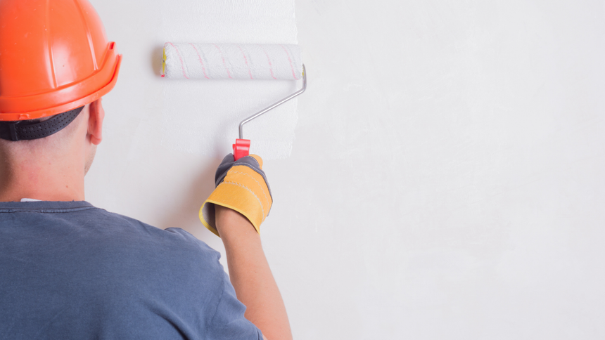 Person wearing a hard hat and gloves painting a white wall with a paint roller.