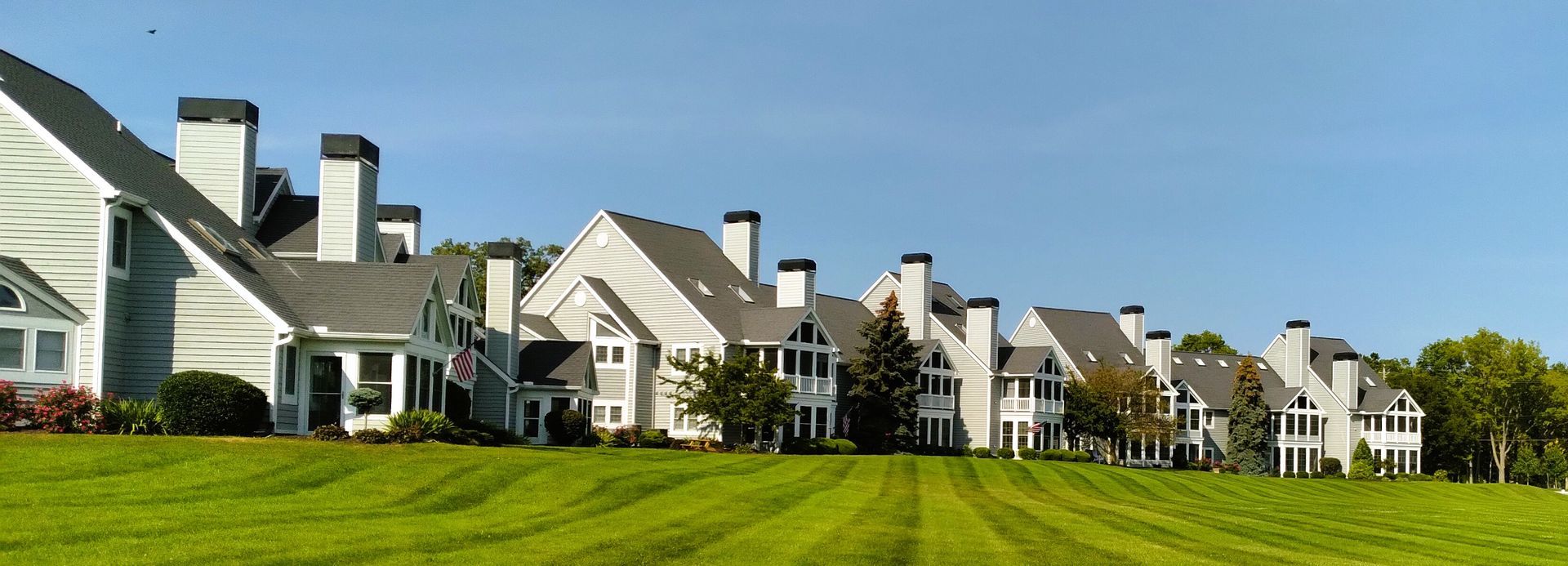 A row of light-colored, multi-story townhomes with dark roofs and chimneys situated above a wide, green grassy lawn.