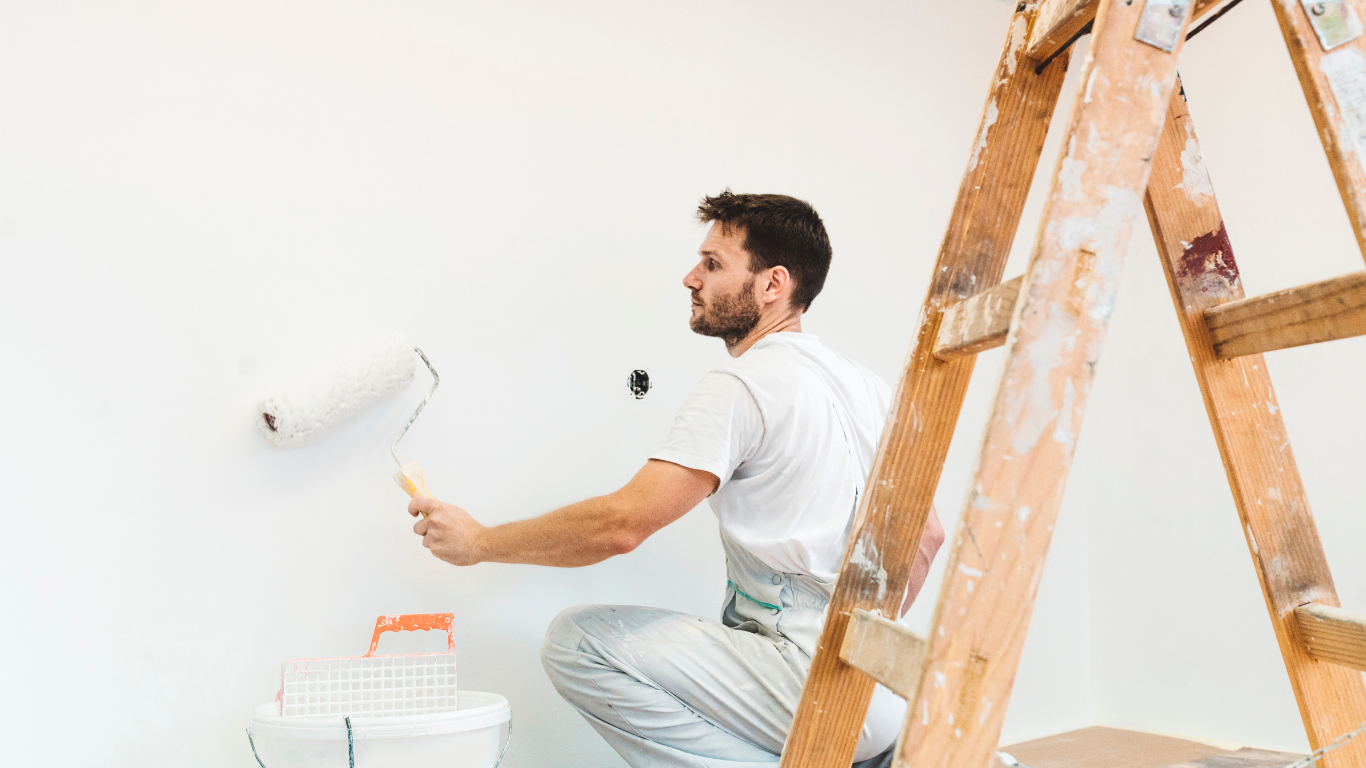Man paints a white wall with a roller while on his knees next to a wooden ladder.