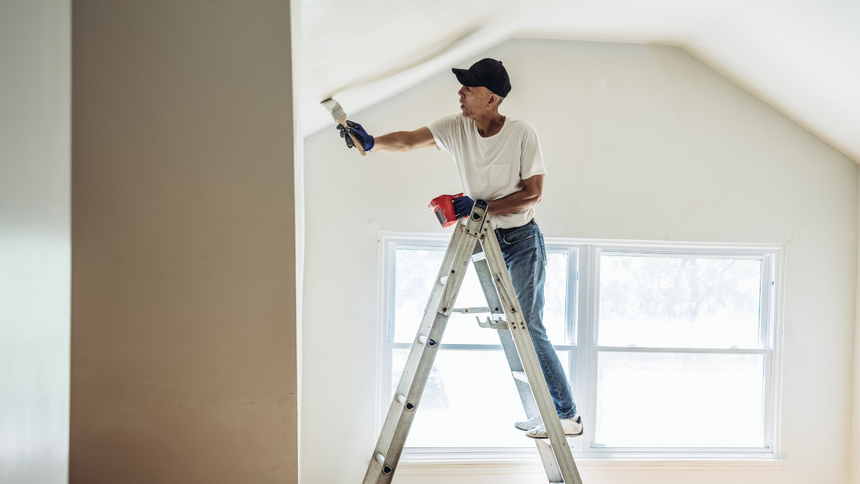 Person painting a white ceiling from a ladder in a room with a window.