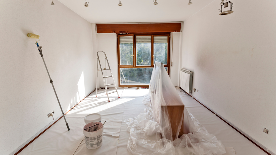 Room being painted, with roller, ladder, window, covered furniture, and white drop cloths.