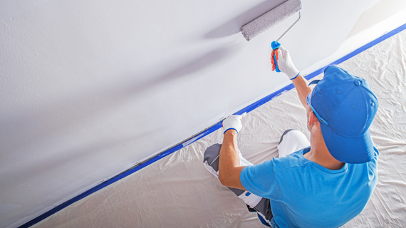 Person painting a white ceiling with a paint roller, using blue painter's tape and wearing a blue cap and gloves.