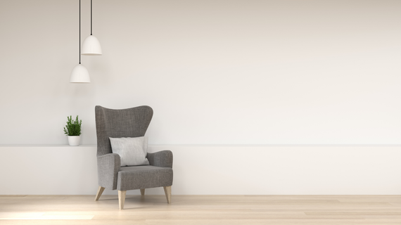 Gray armchair with white pillow, plant, and two pendant lights against a white wall and wood floor.