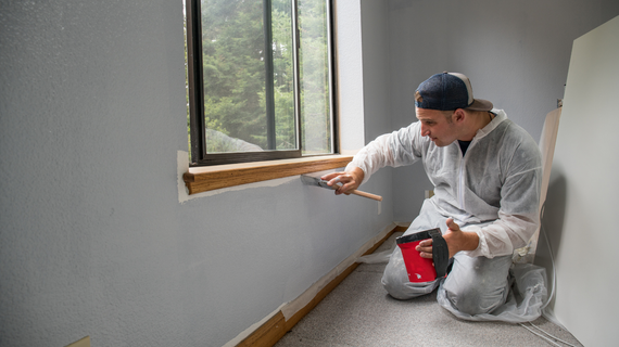 Person in protective suit applying putty to a window frame. Gray wall, wooden trim, and tablet.