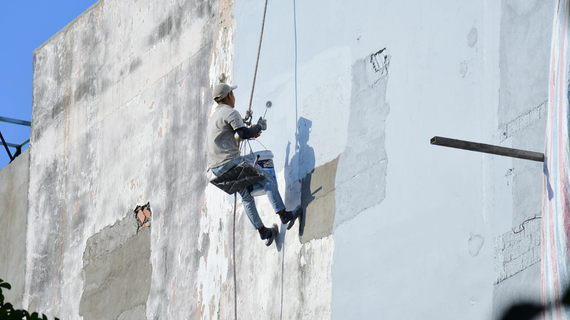 A person painting a white wall while suspended on ropes outdoors.
