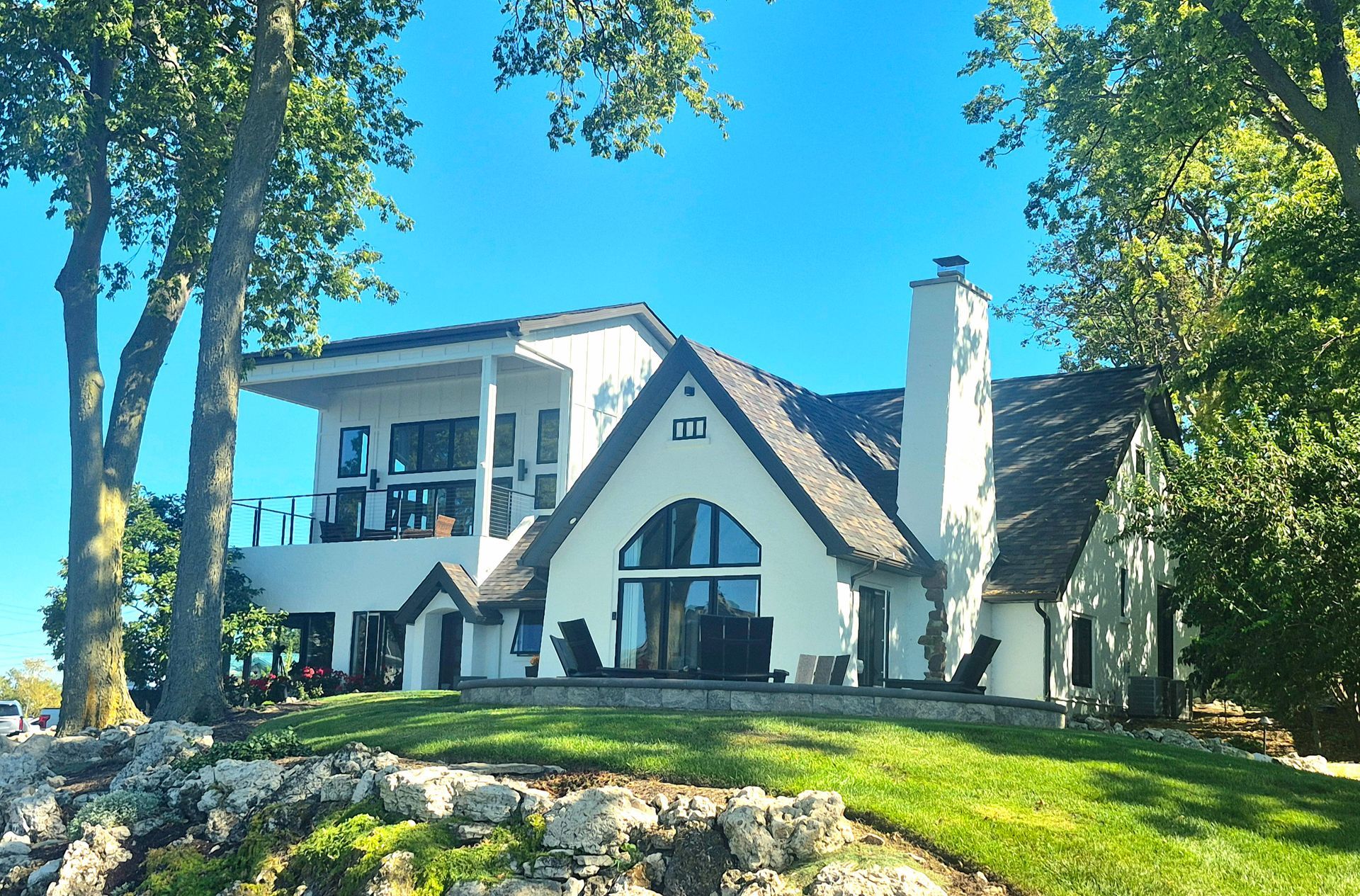 A white, two-story house with a prominent chimney, dark roof, and arched windows, surrounded by trees on a grassy hill.