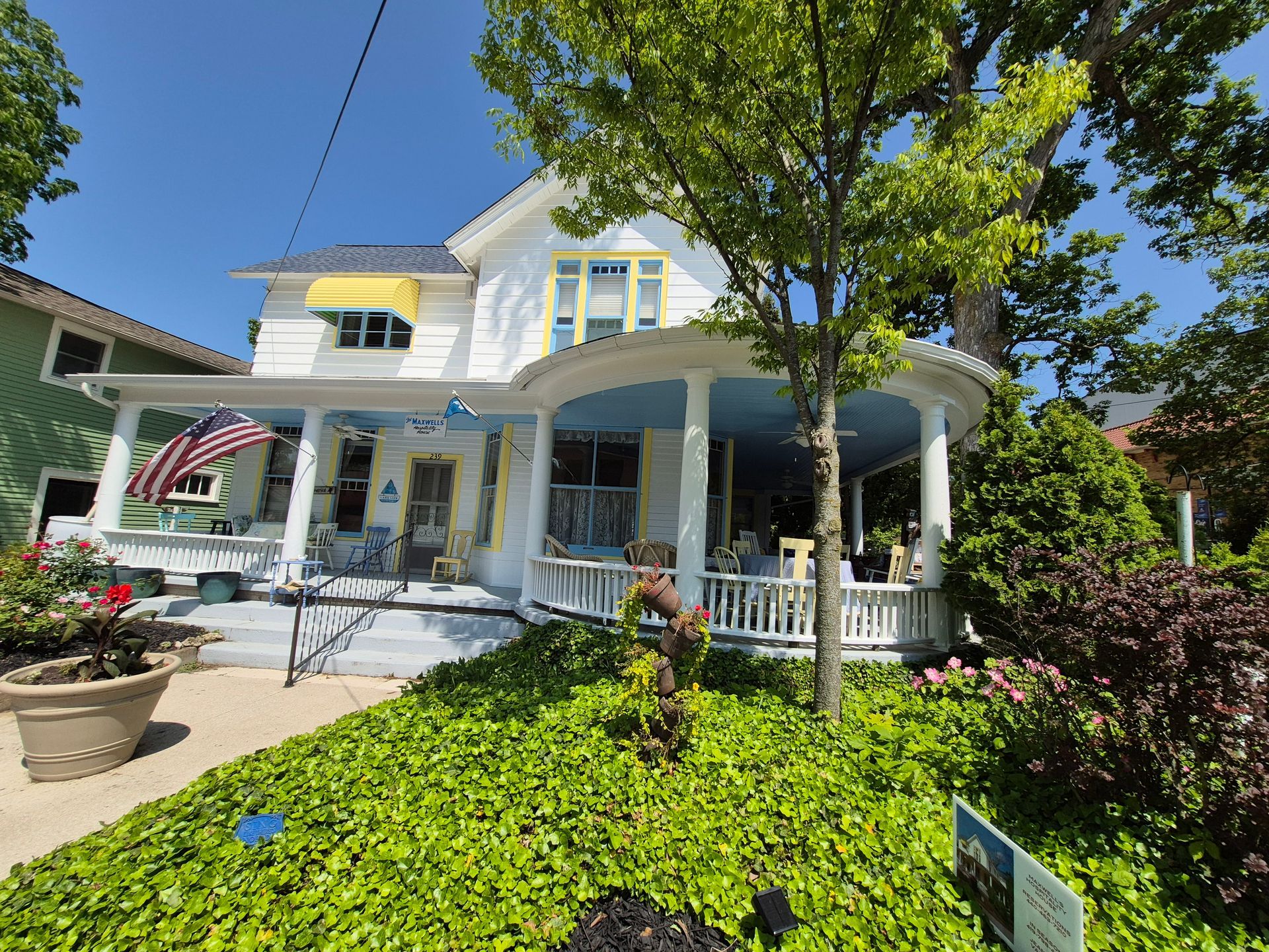 A white two-story house with a blue-ceilinged, wrap-around porch, surrounded by lush green foliage on a sunny day.